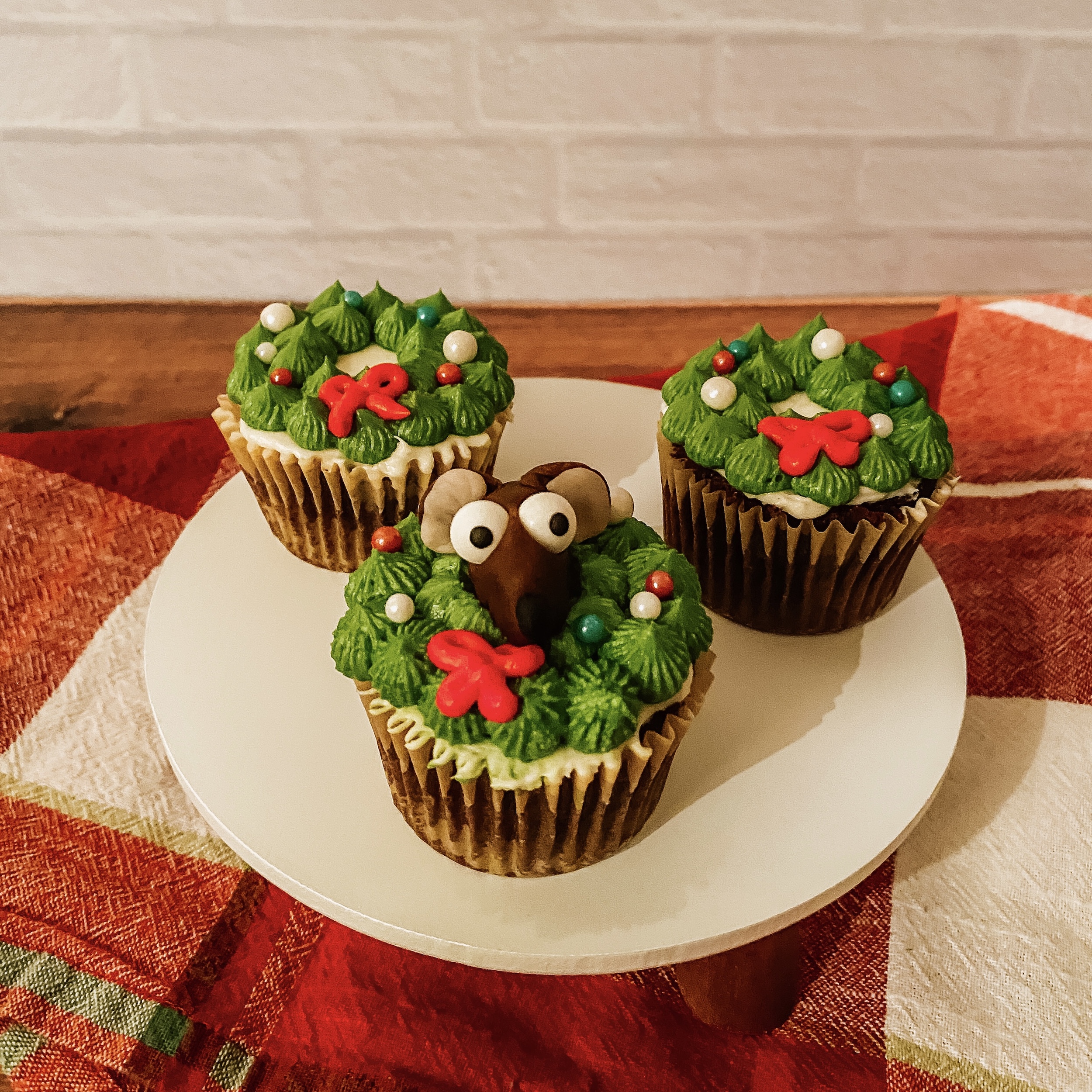 Rizzo's "Here for the Food" Cupcakes (chocolate spice cupcakes with a buttercream christmas wreath and a fondant Rizzo head) on a plate with a red and green plaid napkin beneath it.