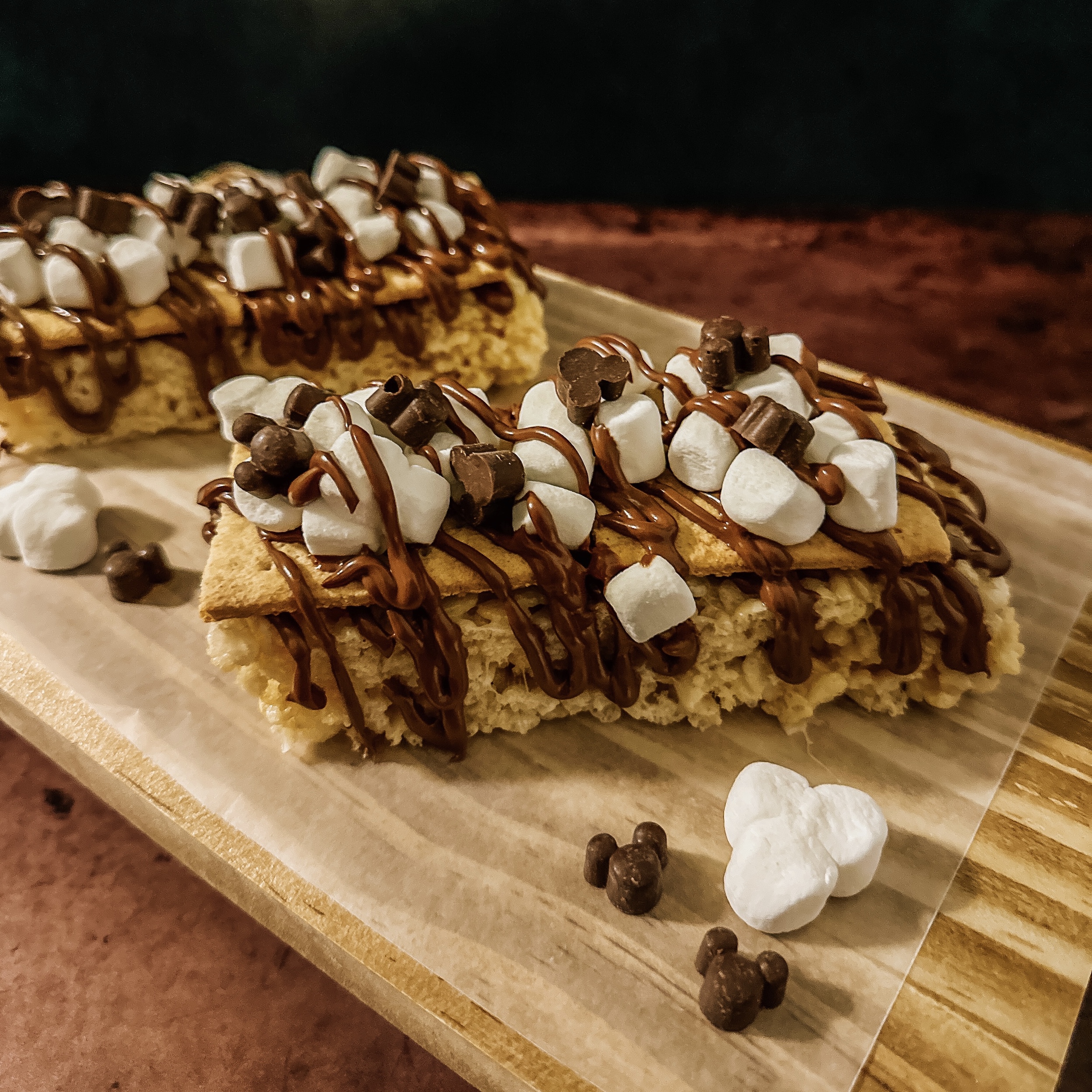 Disney Parks S'mores Cereal Treat Bars (Rice krispies treat with graham cracker, chocolate drizzle, mini marshmallows, and mickey chips on a wooden tray)