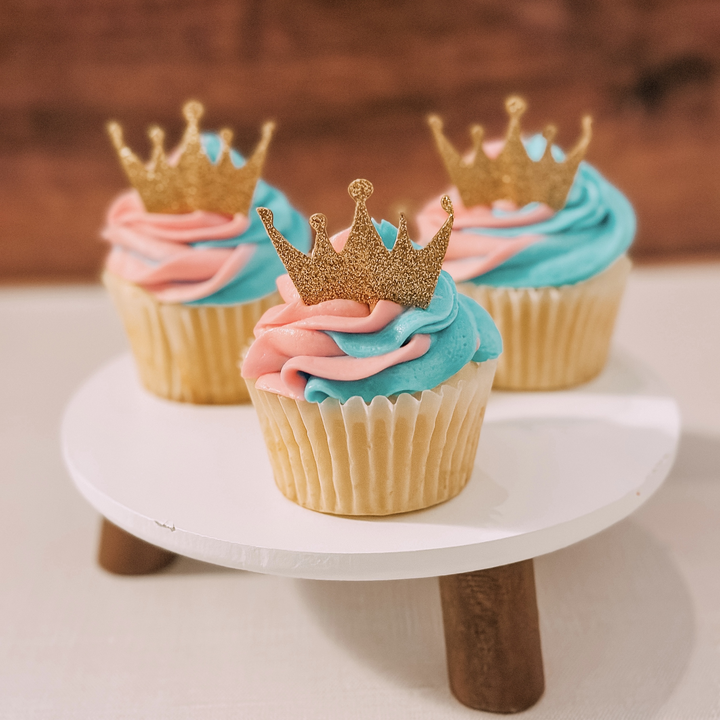Three Sleeping Beauty Cupcakes (vanilla cupcakes with pink and blue swirl frosting and gold crown topper) on a platter